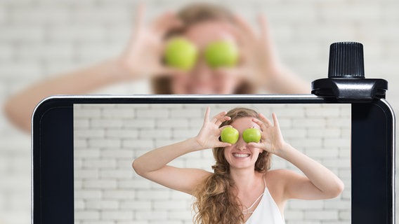 Die Kamera eines Smartphones zeigt eine lachende Frau, die sich zwei Äpfel vor die Augen hält. © colourbox 