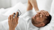 Ein im Bett liegender Mann hält sich mit einer Hand den Kopf, in der anderen ein Smartphone © colourbox 