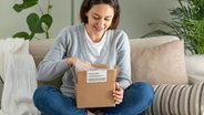 Eine junge Frau sitzt auf einem Sofa und schaut erfreut in ein geöffnetes Paket © colourbox 