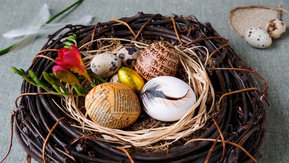 Drei verschieden gestaltete Eier liegen in einem Kranz aus Zweigen, eine rote Blüte liegt obenauf. © NDR Foto: Anja Deuble