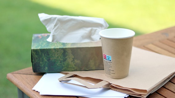 Druckerpapier, Papiertaschentücher, eine Papiertüte und ein Papierbecher auf einem Holztisch vor Rasen im Hintergrund © NDR Foto: Elke Janning