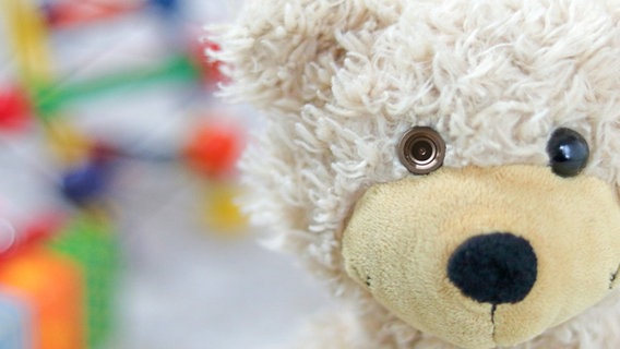 Nahaufnahme eines Teddys, auf dem sein Gesicht zu sehen ist mit einer Kamera im Auge und buntem Spielzeug im Hintergrund © NDR Foto: Elke Janning