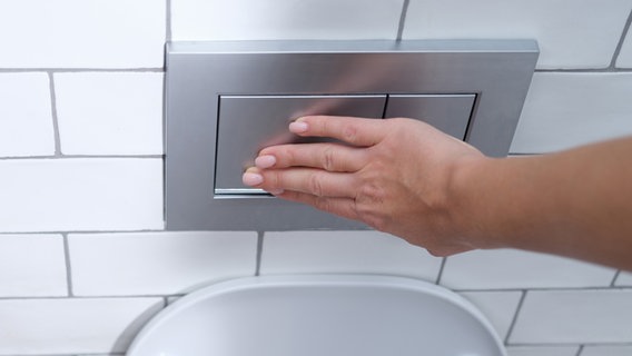 Eine Hand betätigt eine Taste an der  Wasserspülung einer Toilette. © Colourbox 
