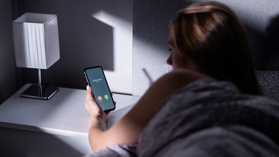 Im Bett liegende Frau schaut auf ihr Smartphone-Display, auf dem ein Unbekannter anruft. © Colourbox.de Foto: Colourbox.de