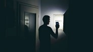 Ein Mann leuchtet mit seinem Smartphone in der dunklen Wohnung und überprüft den Sicherungskasten. © picture alliance / CHROMORANGE Foto: Michael Bihlmayer