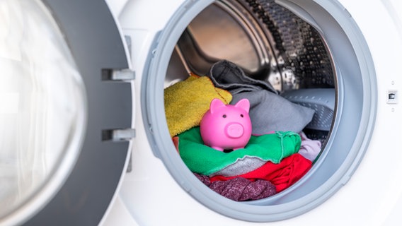 Blick in eine Waschmaschine, in der auf der Wäsche ein Sparschwein steht. © colourbox 