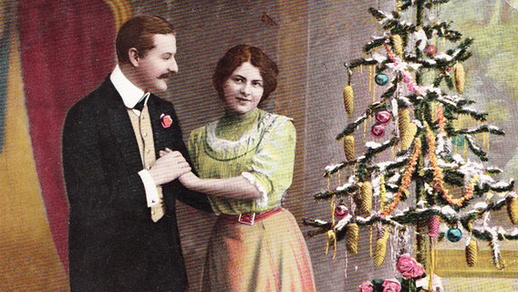 Auf einem historischen Bild etwa von 1910 steht ein Paar neben einem geschmückten Weihnachtsbaum. © fotolia Foto: Patricia