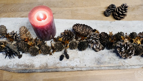 Eine Girlande aus Tannenzapfen liegt auf einem alten Brett, daneben steht eine brennende rote Kerze. © NDR Foto: Anja Deuble