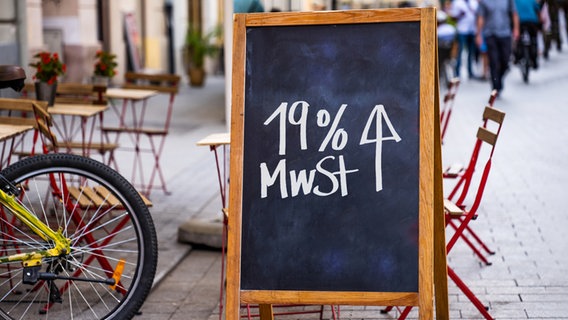 Eine Tafel mit der Aufschrift "19 Prozent Mehrwertsteuer (MwSt) und einem Pfeil, der nach oben zeigt" steht vor leeren Stühlen, Tischen und einem Fahrrad. © Colourbox Foto: Skrypko Ievgen