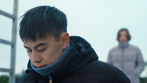 Ein dunkelhaariger Junge in Jacke und Schal blickt mit ernstem Gesichtsausdruck nach unten. © Mothership/NDR 