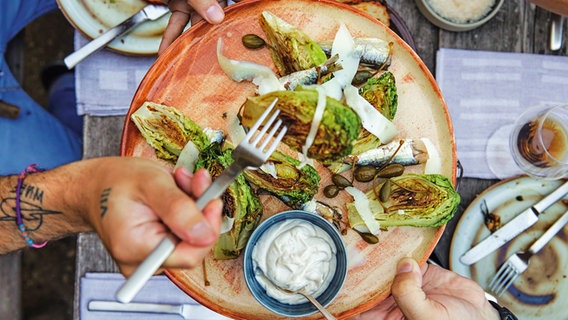 Gegrillte Salatherzen liegen mit einem Dipschälchen auf einem Teller. Eine Männerhand nimmt sich ein Salatherz mit der Gabel. © ZS/Jo Kirchherr Foto: Jo Kirchheer
