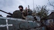 Eine ukrainische Sanitäterin mit Helm und Tarnjacke schaut aus einer Luke eines gepanzerten Mannschaftswagens heraus und hebt die Hand zum Gruß. © picture alliance/dpa/SOPA Images via ZUMA Press Wire Foto: Ashley Chan