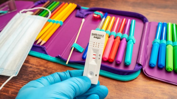 Hände halten einen Covid-19 Schnelltest mit positivem Ergebnis vor einem Etui mit bunten Stiften. © picture alliance/CHROMORANGE/Michael Bihlmayer Foto: Michael Bihlmayer