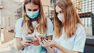 Zwei junge Frauen mit Mundschutz schauen auf ihre Handys. © Photocase Foto: przemekklos
