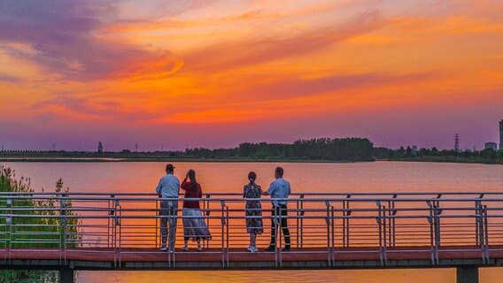 Vier Menschen schauen von einer Brücke in den Sonnenuntergang. © picture alliance / CFOTO | CFOTO Foto: CFOTO