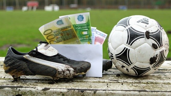 Ein Briefumschlag mit Geldscheinen steckt zwischen FuÃballschuhen, die auf der Auswechselbank eines Sportplatzes stehen. © imago images/Hanno Bode 
