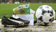 Ein Briefumschlag mit Geldscheinen steckt zwischen FuÃballschuhen, die auf der Auswechselbank eines Sportplatzes stehen. © imago images/Hanno Bode 
