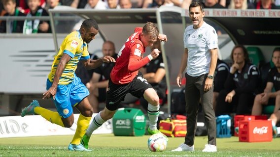 Hannovers Maximilian Beier (Mitte) spielt gegen Braunschweigs Nathan de Medina. Rechts steht Hannovers Trainer Stefan Leitl. © picture alliance/dpa | Swen Pförtner 