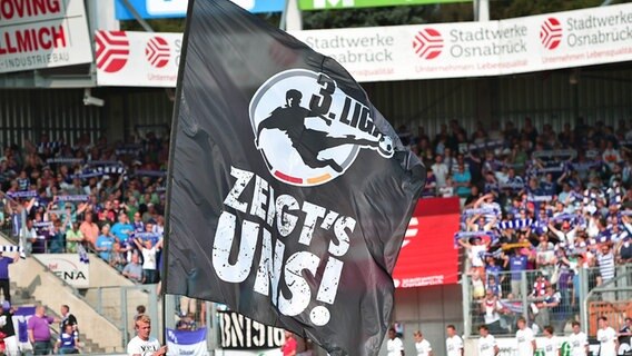 Eine Fahne mit der Aufschrift "Dritte Liga, zeigt's uns" wird im Stadion des VfL Osnabrück geschwenkt. © picture alliance / dpa / Revierfoto 