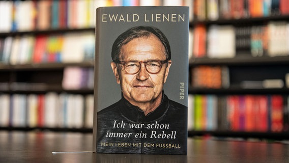 Ewald Lienens Biographie "Ich war schon immer ein Rebell" steht auf einem Tisch vor einem Bücherregal. © Witters 