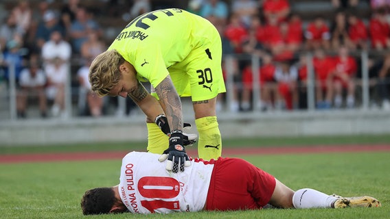 Phönix-Keeper Carl Leonhard beugt sich über den verletzten VfB-Lübeck-Spieler Manuel Farrona Pulido. © IMAGO / Susanne Hübner 