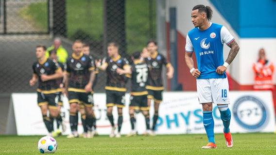 Rostocks Juan Jose Perea ist enttäuscht, während im Hintergrund Karlsruher Spieler einen Treffer bejubeln. © IMAGO / Fotostand 