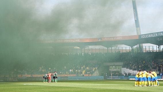 Hannovers und Braunschweigs Spieler stehen jeweils auf dem Rasen zusammen, während Rauchschwaden durch das Stadion ziehen. © picture alliance/dpa | Swen Pförtner 