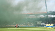 Die Spieler von Hannover und Braunschweig stehen gemeinsam auf dem Rasen, während Rauchwolken durch das Stadion ziehen. © picture Alliance/dpa | Swen Pförtner 
