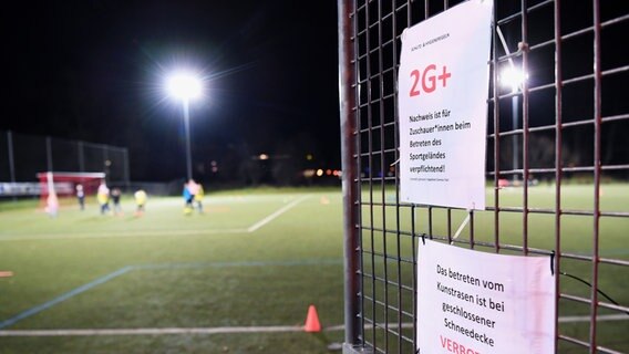 Am Zaun eines Fußballplatzes hängt ein Schild, das darauf hinweist, dass hier für Zuschauer nach der 2G-Plus-Regel verfahren wird. © picture alliance / Pressebildagentur ULMER 