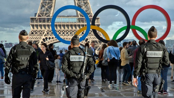 Französische Polizisten patrouillieren in Paris vor dem Eiffelturm, wo die Olympischen Ringe stehen. © picture alliance / Michel Euler/AP/dpa | Michel Euler 