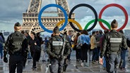 Französische Polizisten patrouillieren in Paris vor dem Eiffelturm, wo die Olympischen Ringe stehen. © picture alliance / Michel Euler/AP/dpa | Michel Euler 