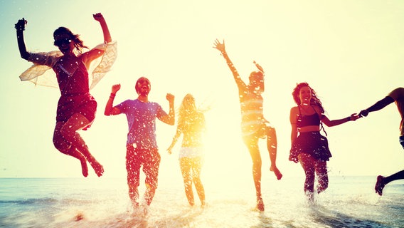 Junge Leute laufen und springen ausgelassen bei einem Sonnenuntergang am Strand aus dem Meer. © fotolia.com Foto: Rawpixel.com