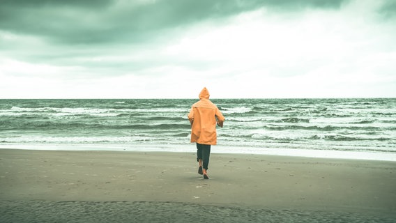 Ein Mensch mit orangefarbener Regenjacke geht am Wasser spazieren. © Photocase Foto: David-W