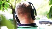 Ein junger Mann sitzt im Grünen und hört Podcasts. Zu sehen ist nur sein Hinterkopf mit den aufgesetzten Kopfhörern. © NDR Foto: Jonas Jung