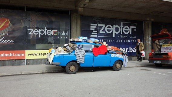 Ein altes hellblaues Auto parkt am Straßenrand, viele Kleidungsstücke sind auf dem Auto abgelegt. © NDR Foto: Maja Bahtijarevic