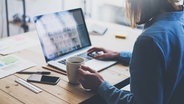 Eine Frau sitzt mit einer Tasse Kaffee an einem Arbeitstisch vor einem Laptop. © fotolia.com Foto: kantver