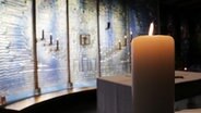 Eine Kerze brennt vor einer blau, weißen Glasscheibe in einer Kapelle. © NDR Foto: Bruno Terenzi Neumann