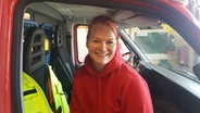 Sonja Haack mit roten Haaren und einem roten Pullover sitzt auf dem Beifahrersitz eines Feuerwehrautos. © NDR Foto: Cassandra Arden