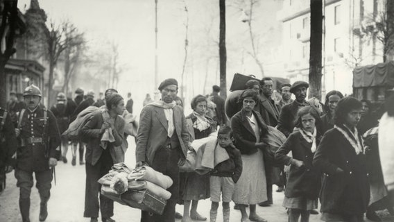 Flüchtlinge stehen mit Gepäck auf einem Gehweg. Neben ihnen laufen Soldaten. © picture alliance / akg-images | akg-images 