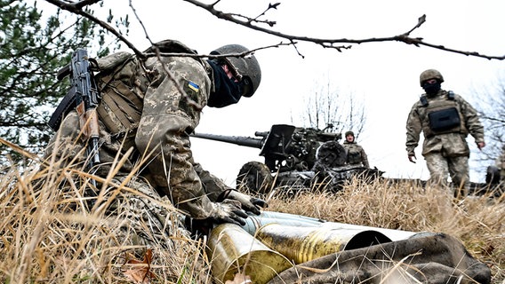 Ein ukrainischer Soldat kniet vor Munition für eine Artilleriegeschütz, welches man im Hintergrund sieht. © IMAGO / Ukrinform Foto: Dmytro Smoliyenko