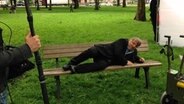 Axel Prahl liegt für einen Dreh auf einer Parkbank © NDR Foto: Patricia Poelk