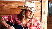 US-Sänger Kid Rock in Karohemd sitzt mit Cowboyhut und Gitarre auf einem Strohballen (Pressefoto von 2010) © Clay Patrick McBride 