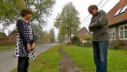 Eine Frau und ein Mann stehen neben einer Dorfstraße an einem vergrabenen Kabel. © NDR 