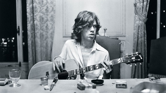 Mick Jagger sitzt mit einer Gitarre in der Hand an einem Tisch. Mick Jagger sitzt mit einer Gitarre in der Hand an einem Tisch. Foto: z90