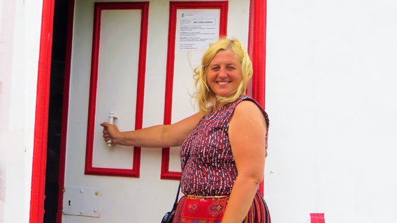 Eine blonde Frau, etwa 50,  mit roter Bluse und rotem Rock offnet eine weiße Holztür. Sie strahlt in die Kamera. © Julia Schulz 