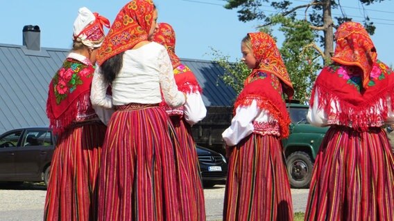 Fünf Frauen mit roten Kopftüchern und Schultertüchern in Trachten stehen auf einem Bauernhof. © Julia Schulz 