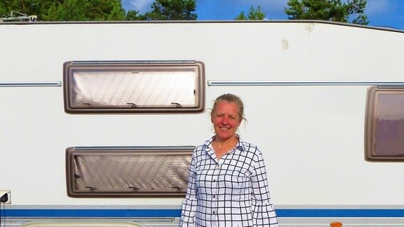 Eine Frau mit Pferdeschwanz steht vor einem Wohnwagen. Sie trägt eine karierte Bluse und lacht in die Kamera. © Julia Schulz 