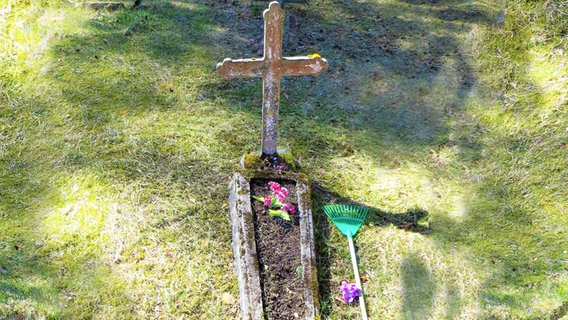 Ein Grab mitten auf einer Wiese. Ein Steinkreuz an dessen Kopfende, darauf steht ein Blumentopf. Neben dem Grab liegt eine Harke. © Julia Schulz 