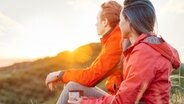 Ein junges Paar mit roten Jacken sitzen auf dem Boden und schauen in einen Sonnenuntergang © Colourbox Foto: Poprotskiy Alexey