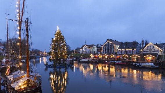 Blich auf einen weihnachtlichen, norddeutschen Hafen mit einem beleuchteten Tannenbaum auf dem Wasser. © imago-images Foto: westend61